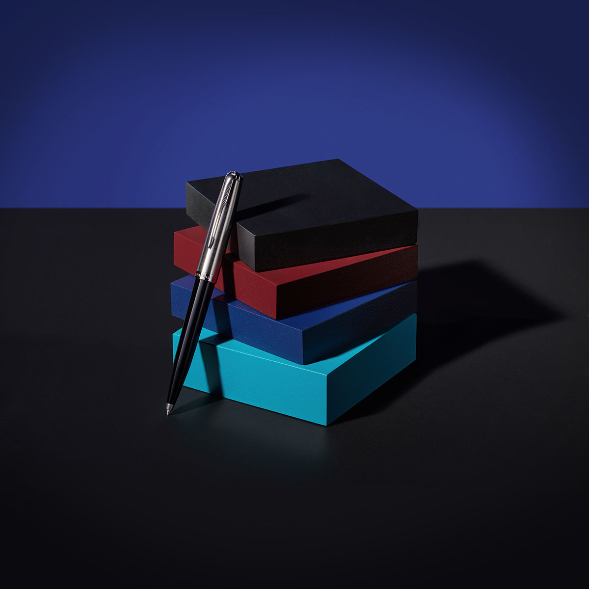 Elegancki długopis Parker oparty o stos kolorowych pudełek w odcieniach czerni, czerwieni, granatu i turkusu.